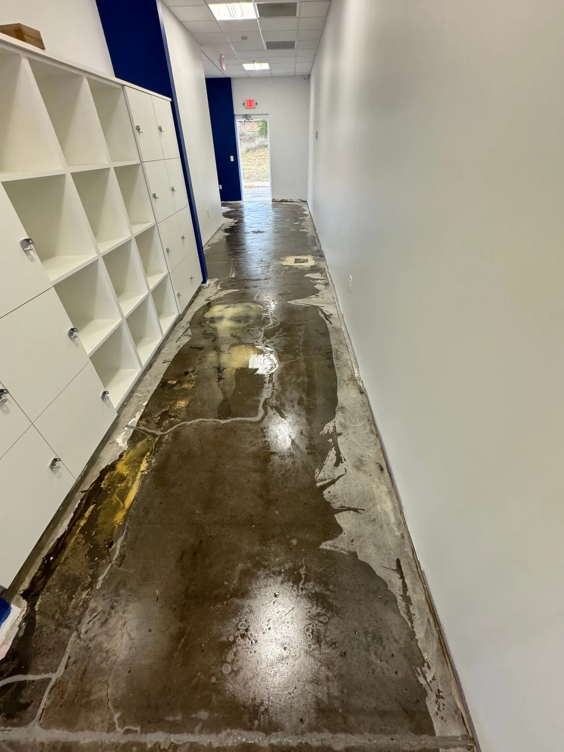 Hallway with damaged, wet floor; lockers on left, white walls, door at end.