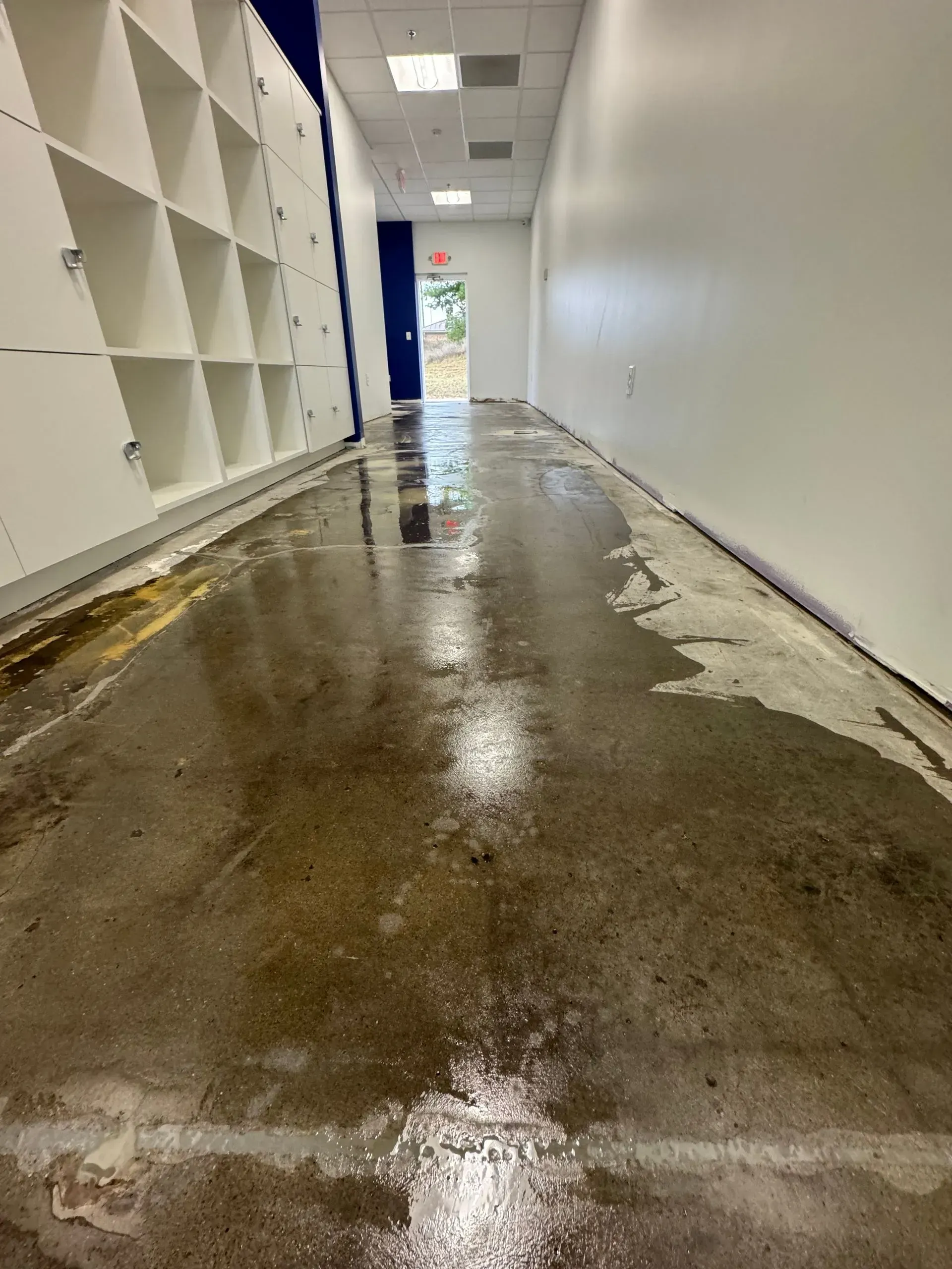 Wet concrete hallway, white walls, lockers, and an open doorway visible at the end.