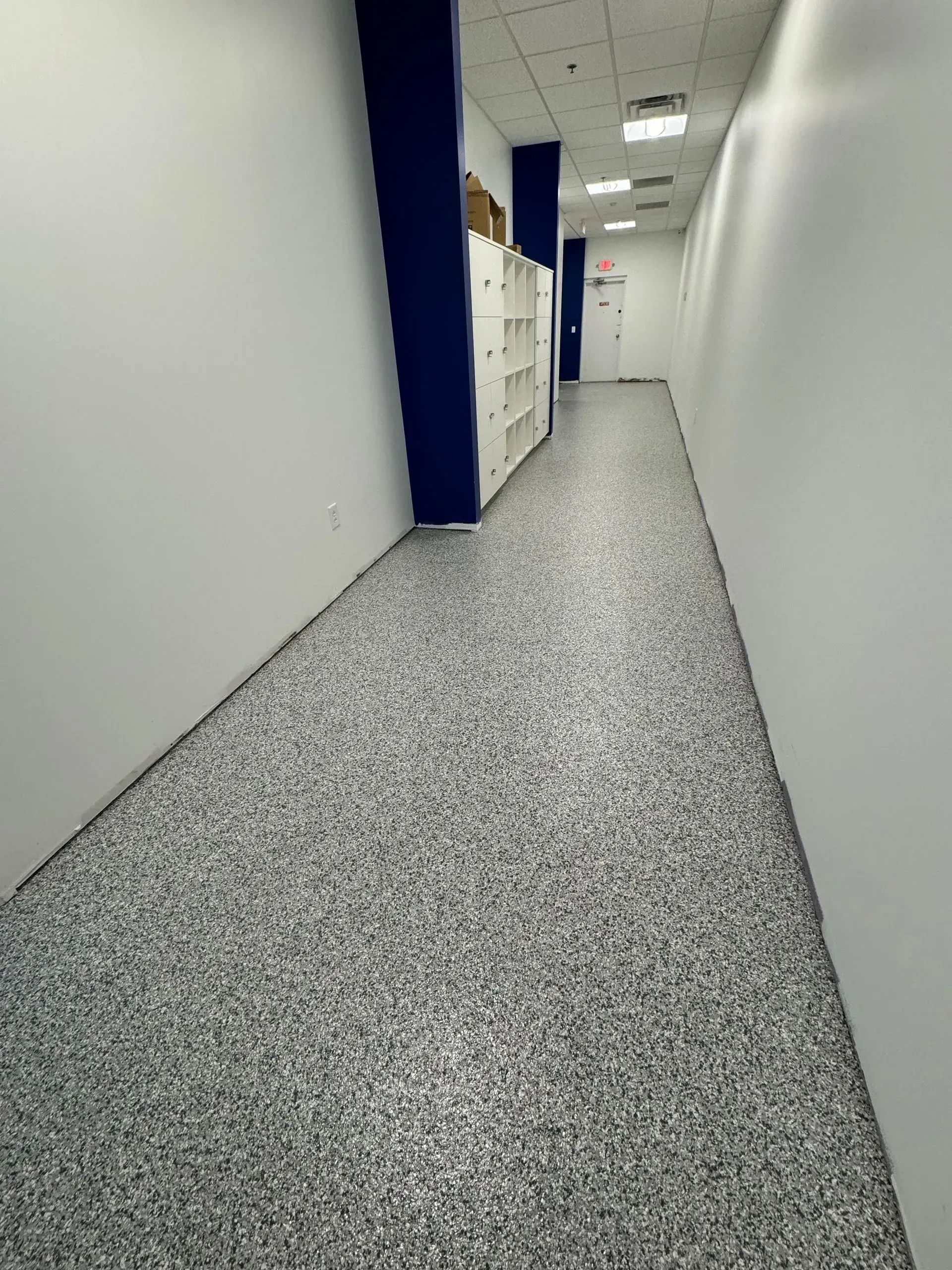 Long hallway with speckled gray floor, white walls, and lockers on the left.
