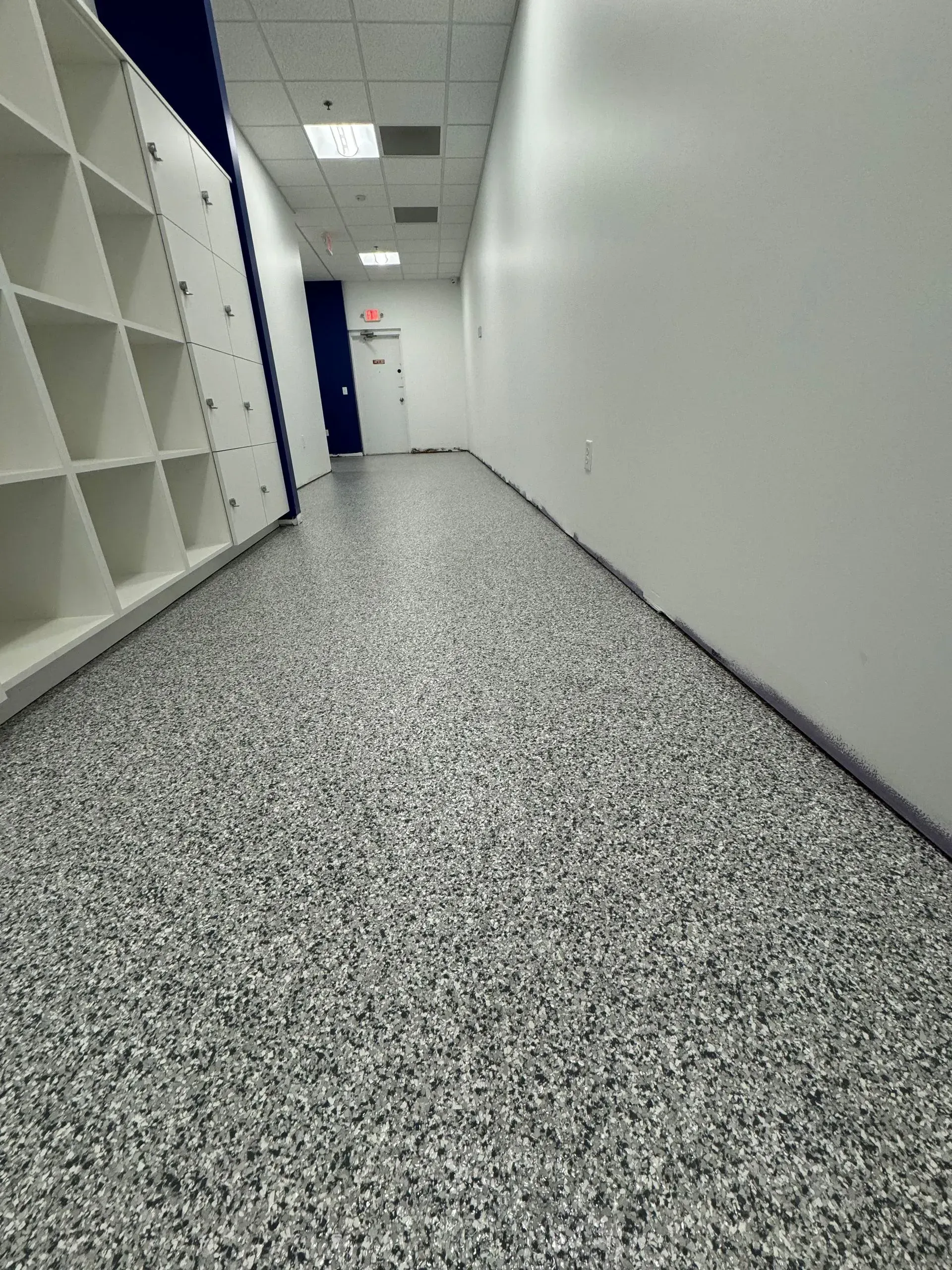 Hallway with gray speckled flooring, white walls, and cubby shelving on the left. A blue door is at the end.