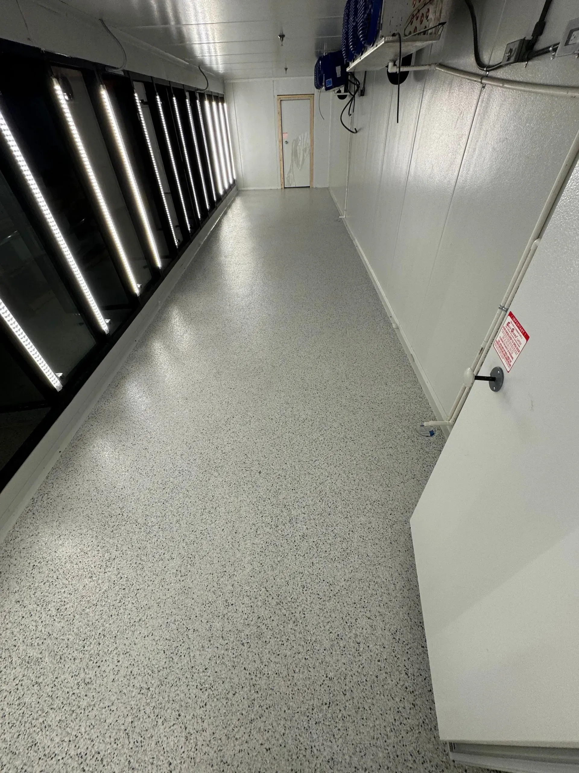 Long narrow room with speckled gray floor, bright lights on the left, a door at the end, and white walls.