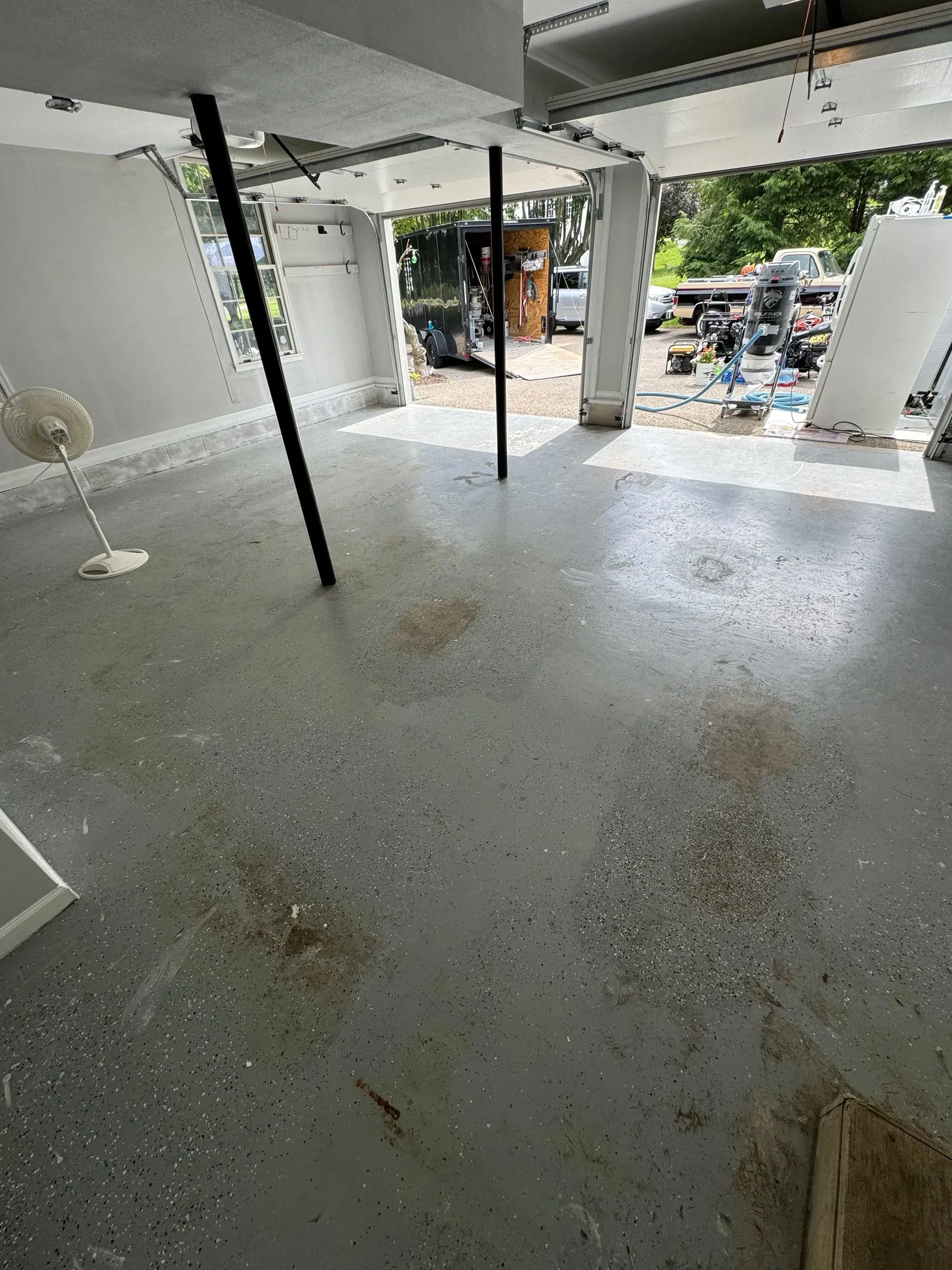 Garage interior with speckled gray floor, support beams, and open garage door revealing outdoor debris.
