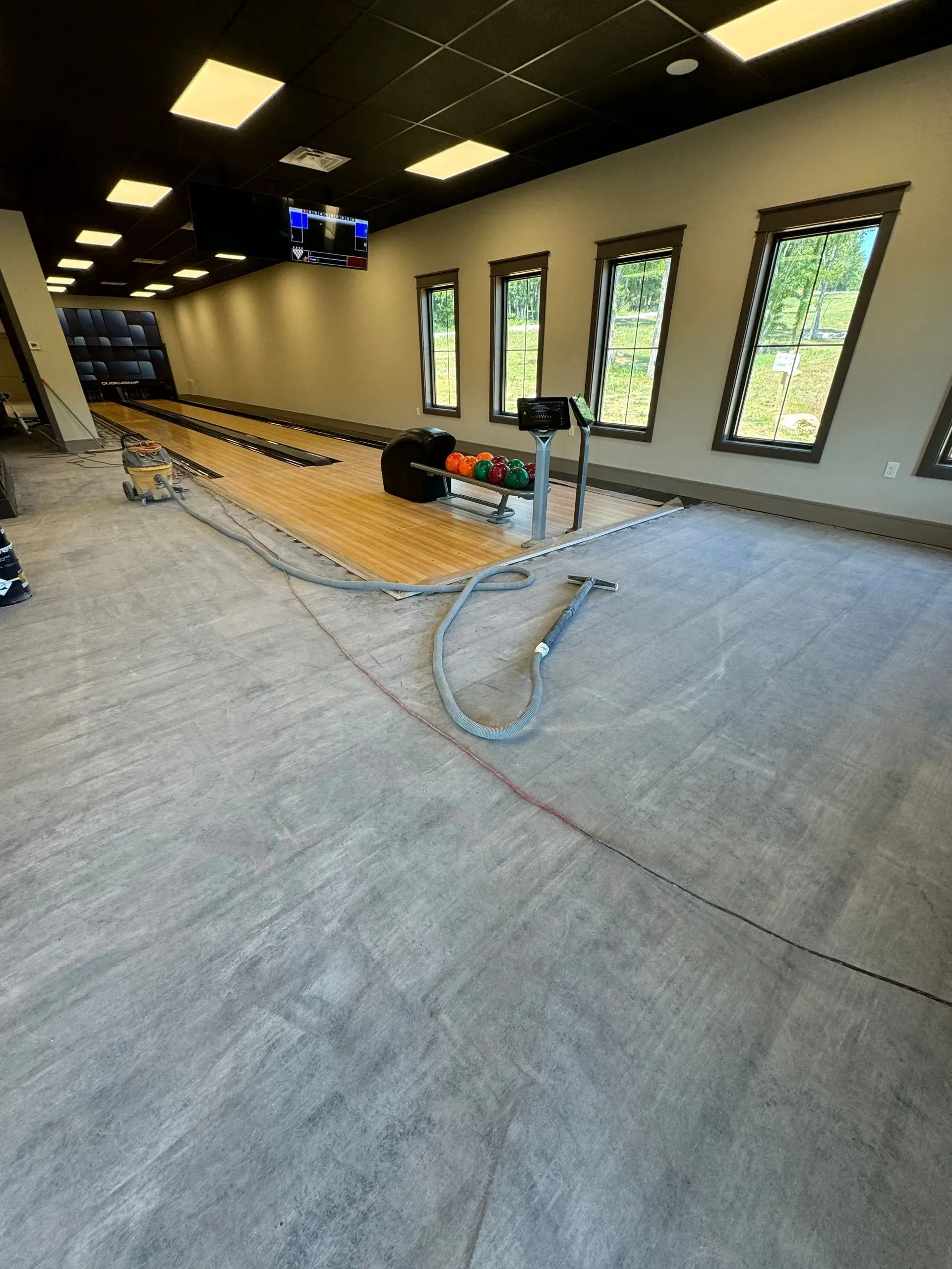 Bowling alley under construction; grey floors, wood lane, windows, equipment, and ceiling lights.