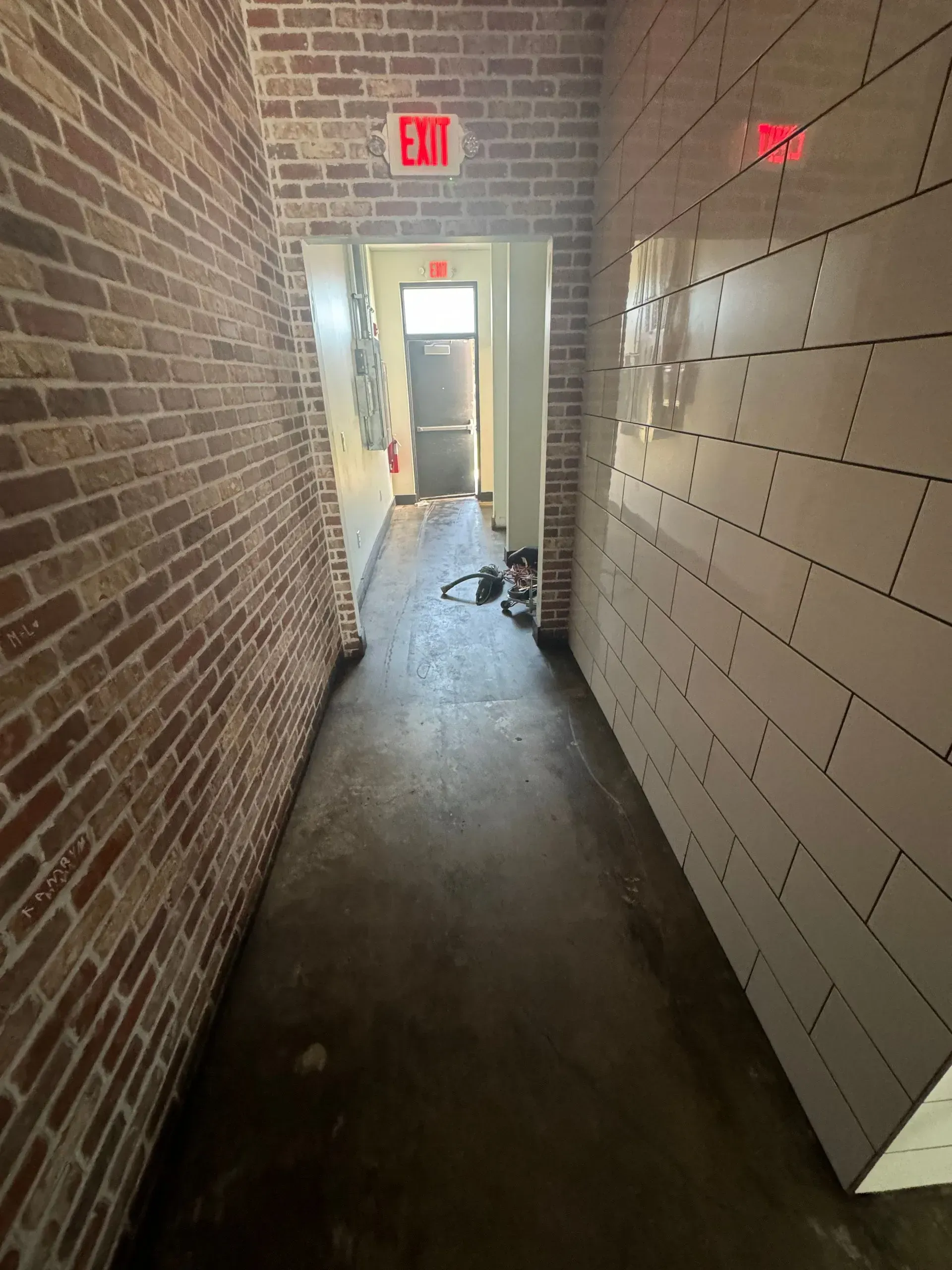 Narrow hallway with brick and tiled walls, exit signs, and a dark floor.