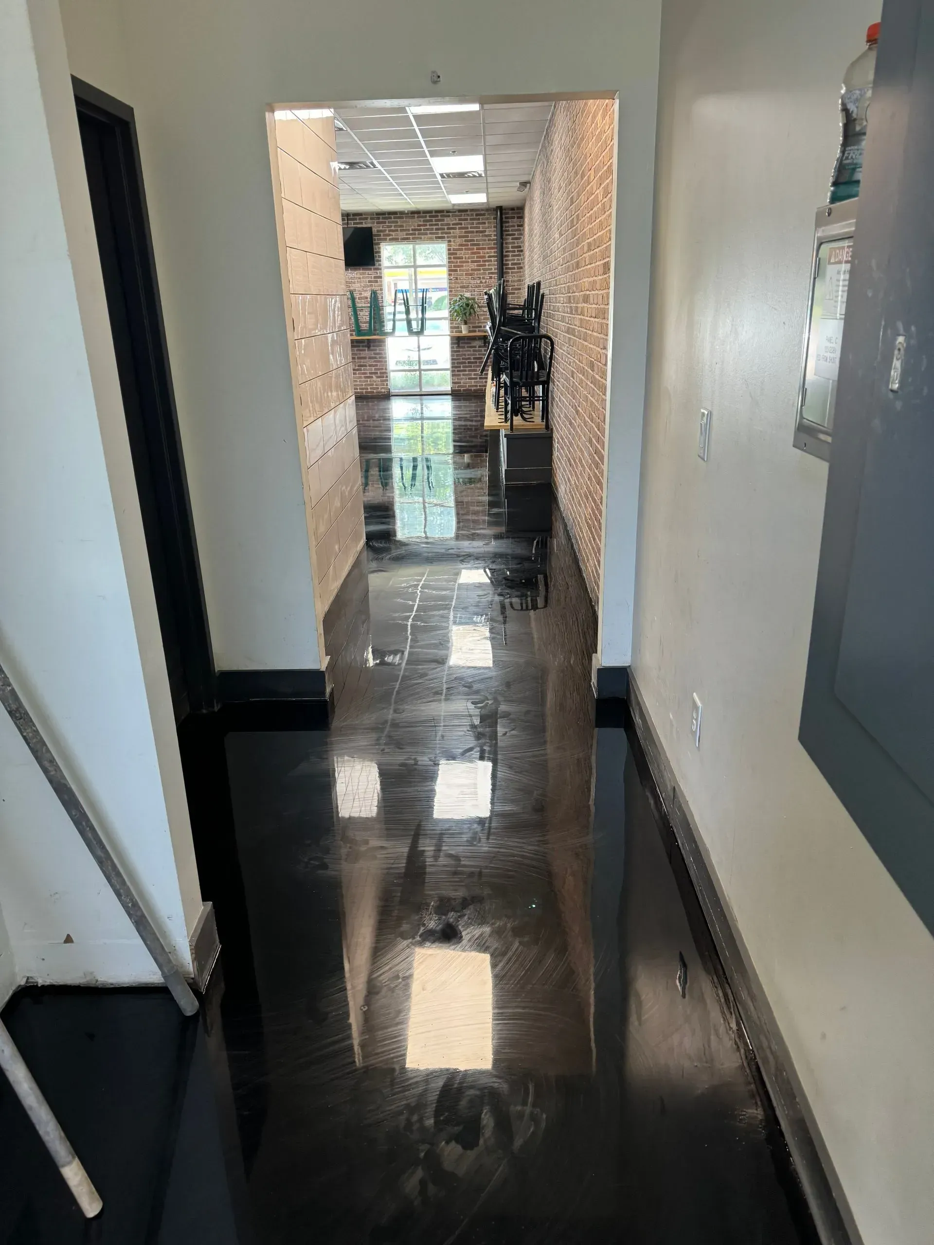 Dark shiny floor in a hallway with an opening to a brightly lit room. Walls are white and brick.