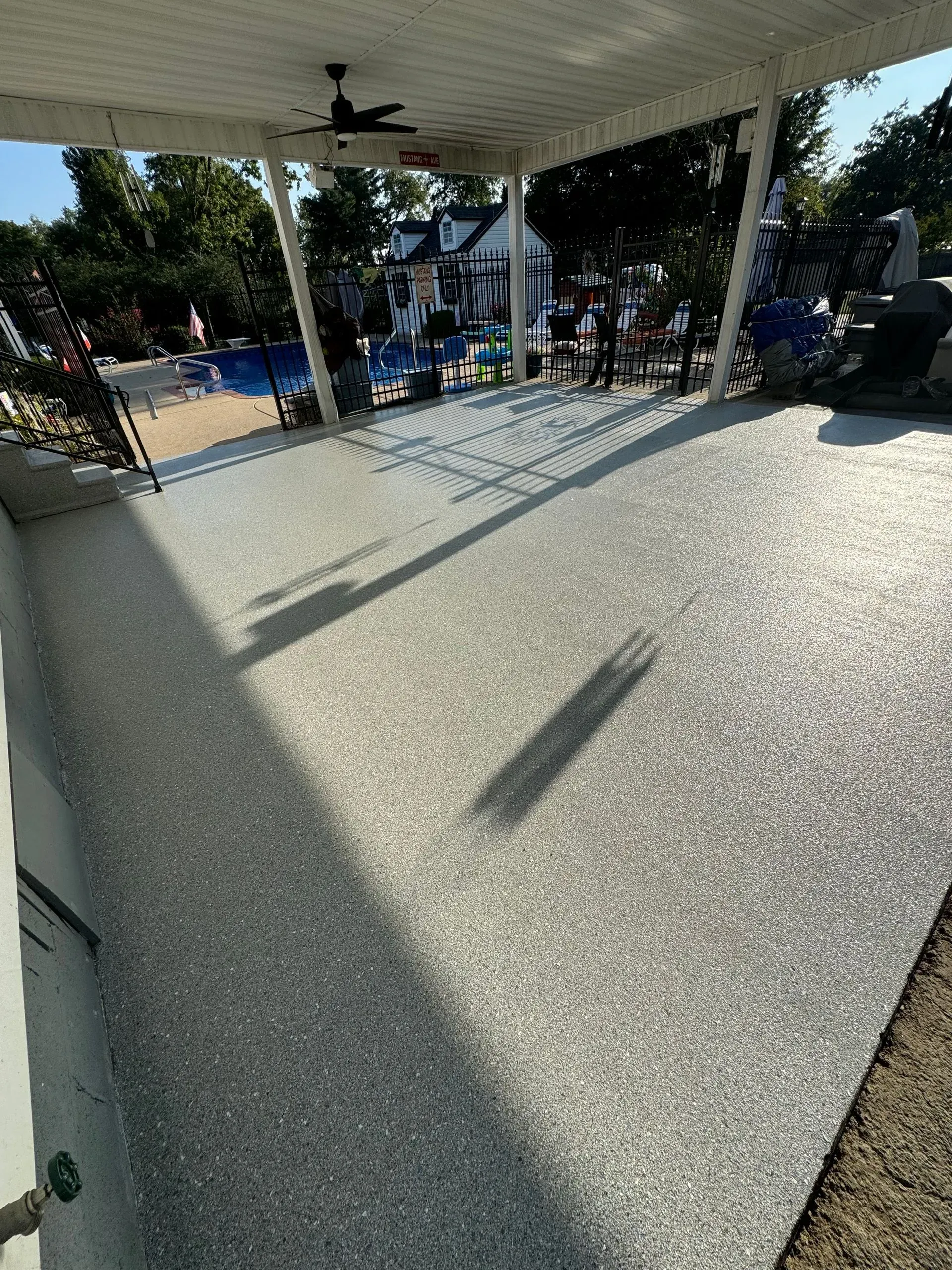 Covered patio with speckled, light gray flooring overlooking a swimming pool and backyard on a sunny day.