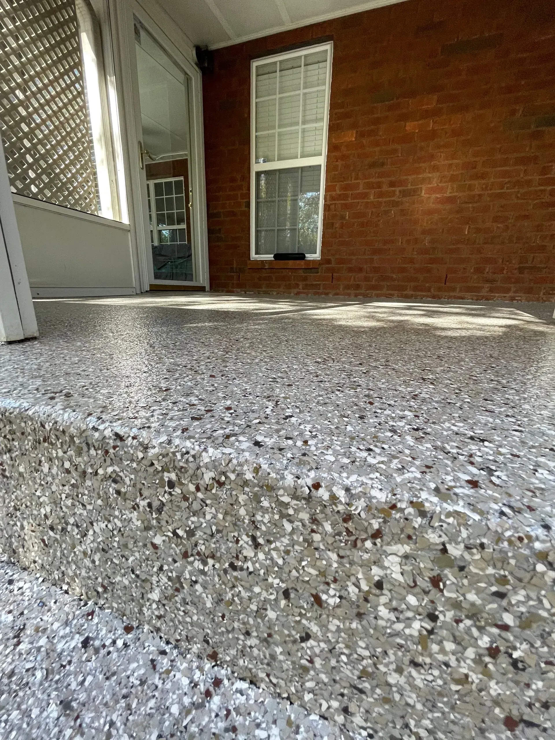 Close-up of a speckled, shiny gray floor on a porch, with a brick wall and window in the background.