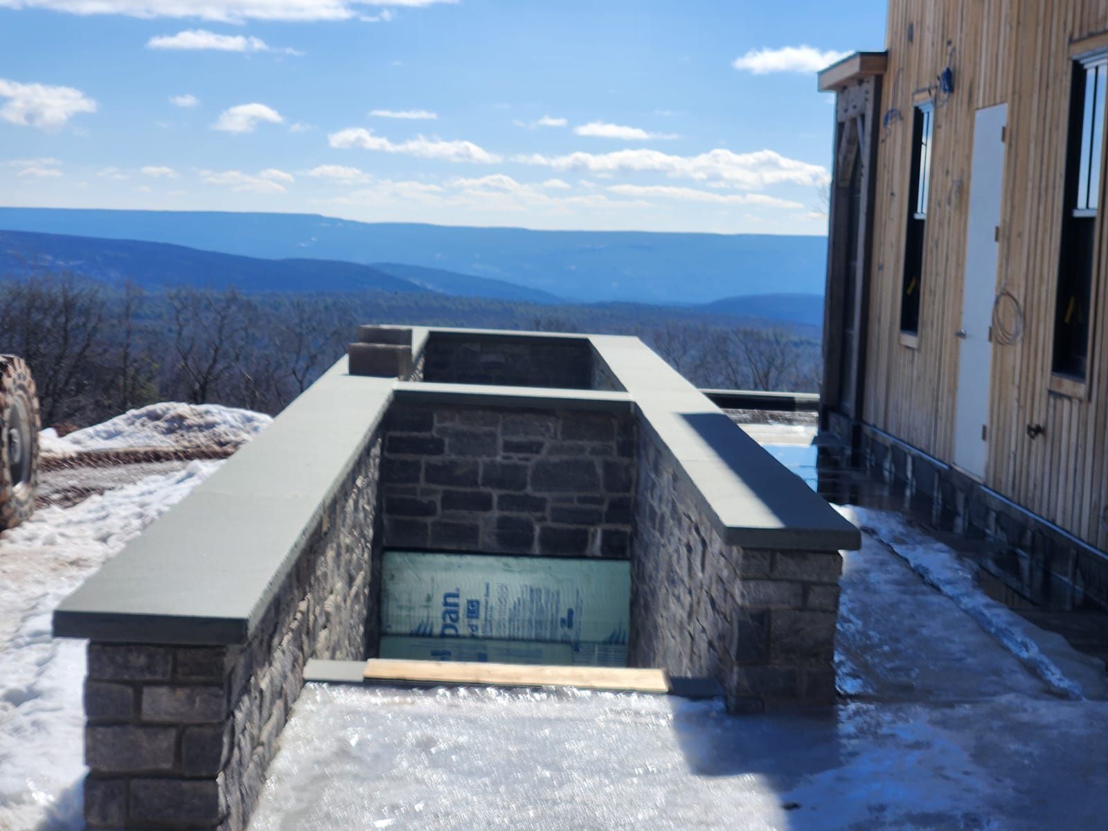 Stone wall and flat concrete walkway overlooking a mountain range. Building on the right. Blue sky.