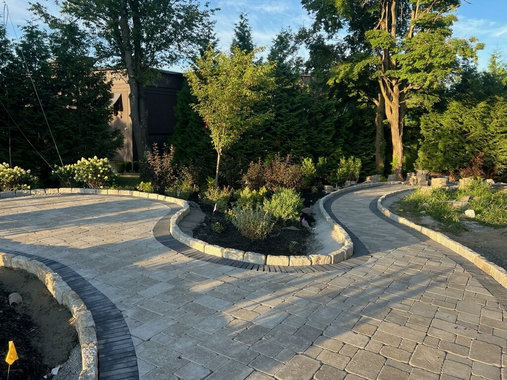 Stone paver driveway with landscaped islands, bordered by dark stone, surrounded by trees.