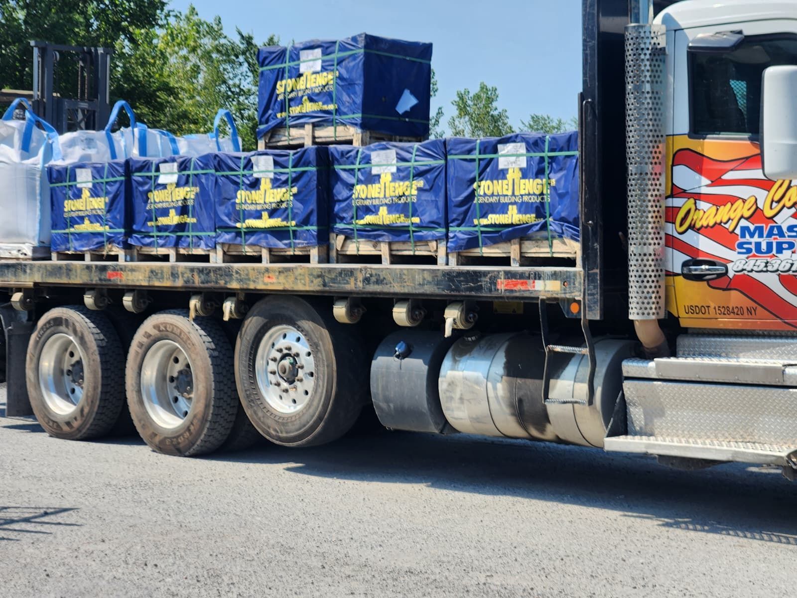 Semi-truck loaded with blue-wrapped pallets, likely construction supplies, parked outdoors. Truck has 
