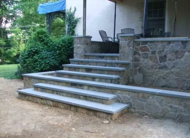 Stone steps leading up to a house with a porch. Natural stone walls and gray steps.