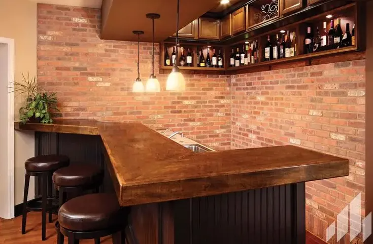 L-shaped bar with brick wall backdrop, brown countertop, shelves of liquor, and stools.