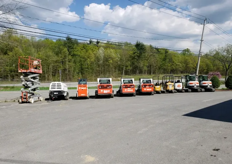 Vehicles lined up on asphalt; includes aerial lift, Bobcat skid steers, and excavators, under a cloudy sky.
