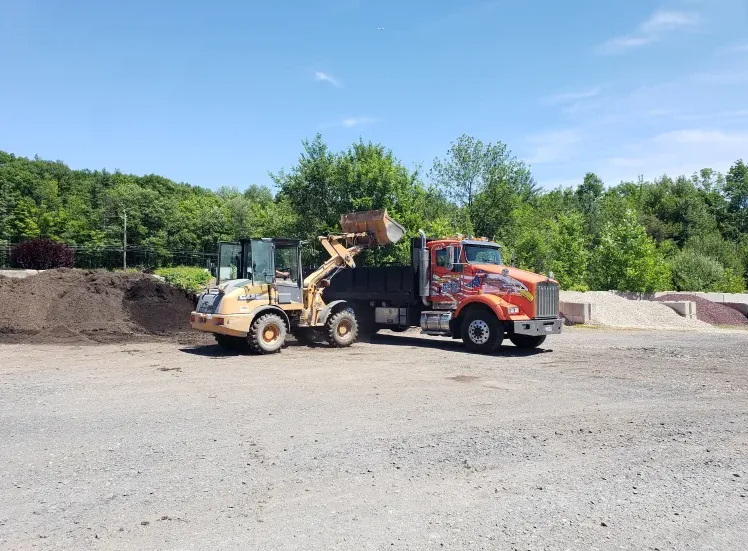 Yellow loader filling orange dump truck with dark material at a gravel yard under a blue sky.