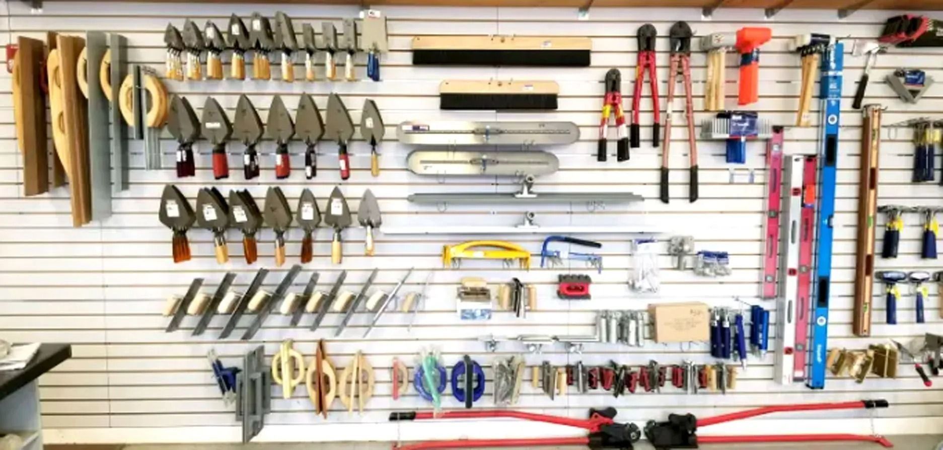 Tools displayed on a white slat wall in a hardware store. Many trowels, levels, and cutters hang.