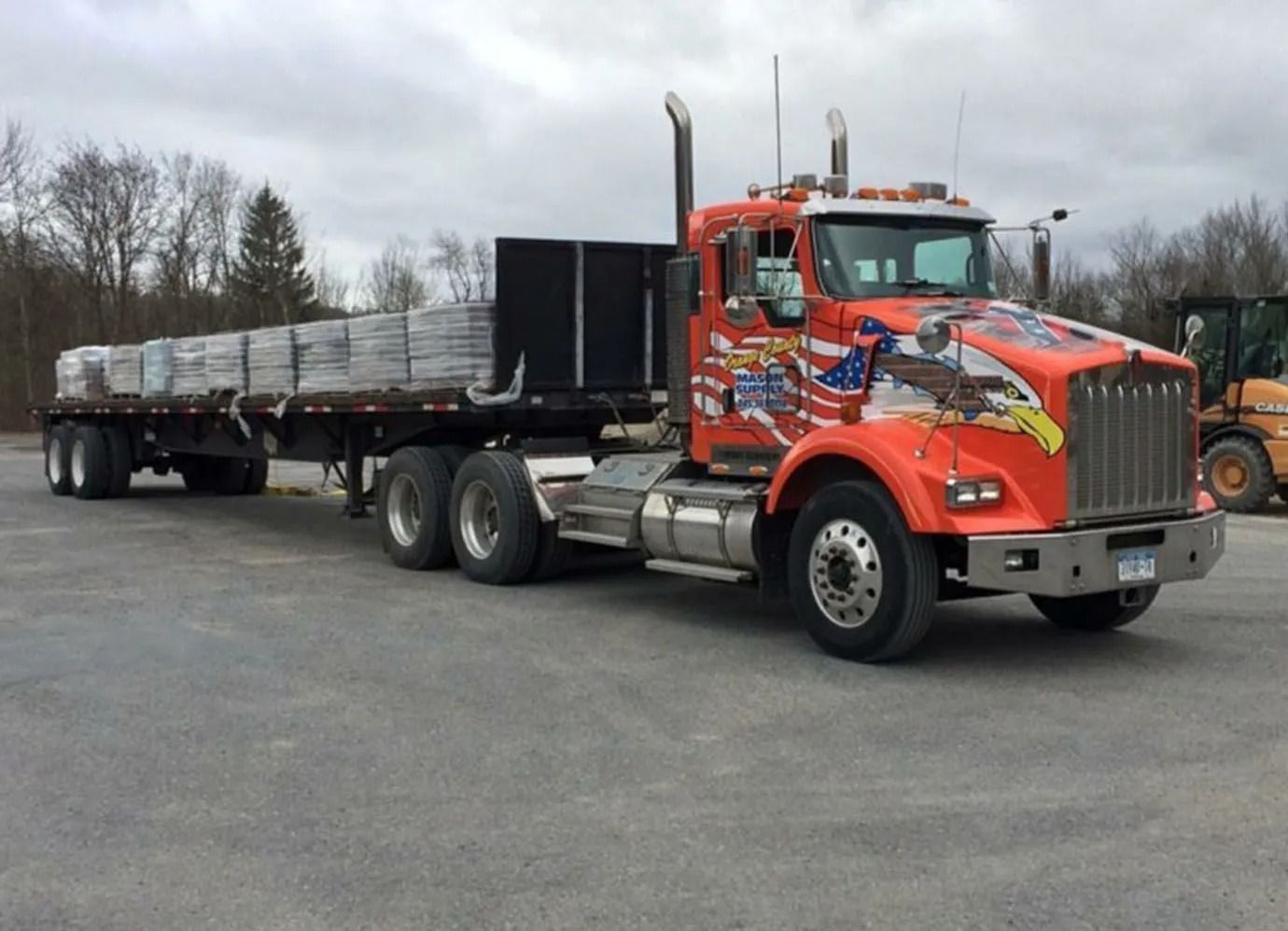 Orange Kenworth flatbed truck hauling a load of materials, parked on asphalt. Eagle graphic.