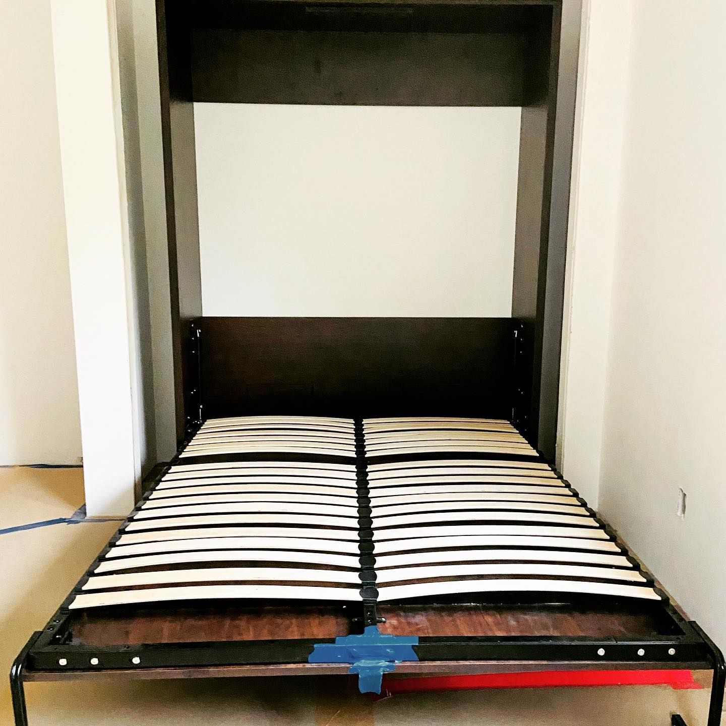 Wall-mounted, dark brown bed frame with a slatted base, lowered. The wall is dark, with a white top panel.