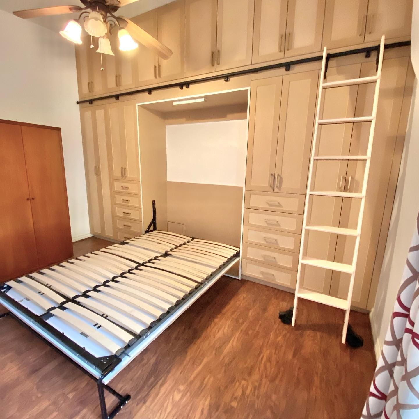 A Murphy bed is partially folded down in a room with built-in storage, a ladder, and a wooden floor.