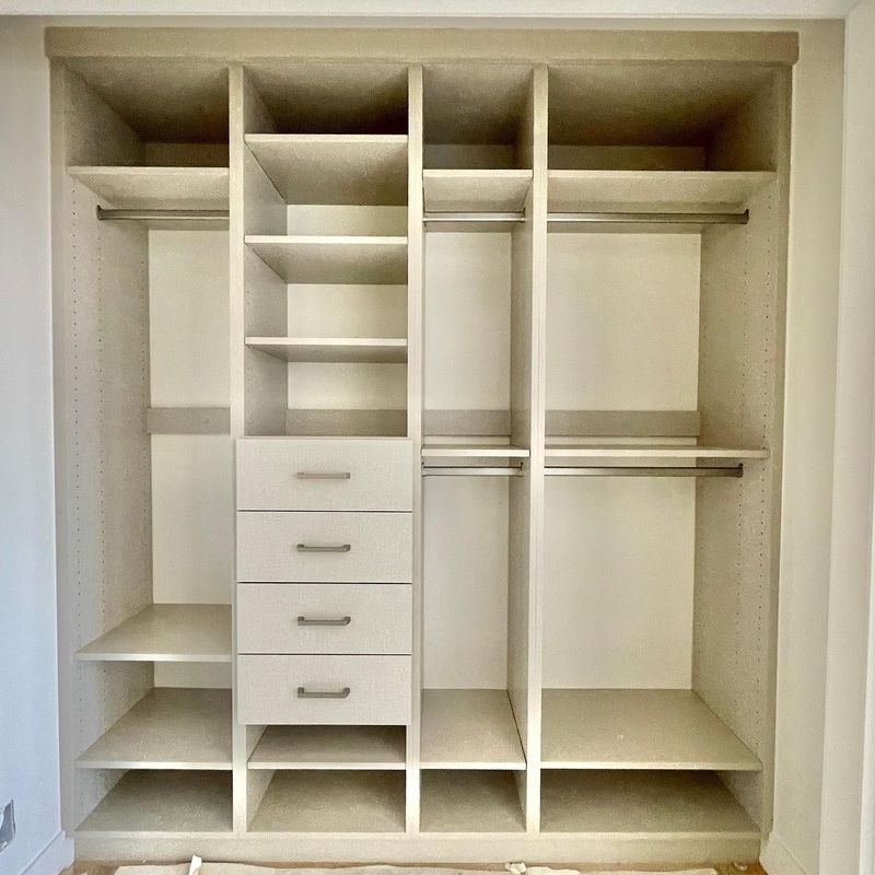 Built-in closet with neutral shelves, drawers, and hanging rods, with light beige interiors.