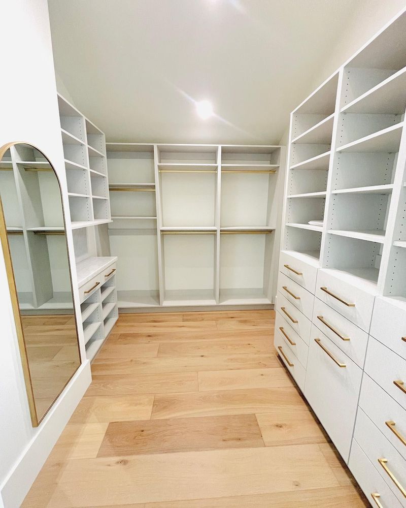 Walk-in closet with white built-in shelving, drawers, and hardwood floors. A gold-framed mirror is on the left.
