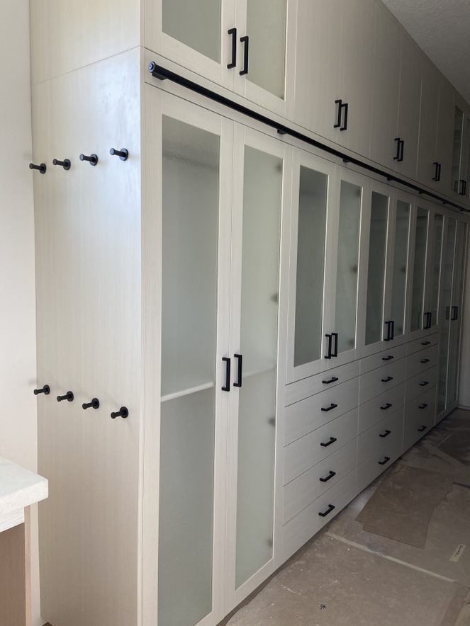 Built-in white closet with glass doors and black handles, drawers, and rods in a room under construction.