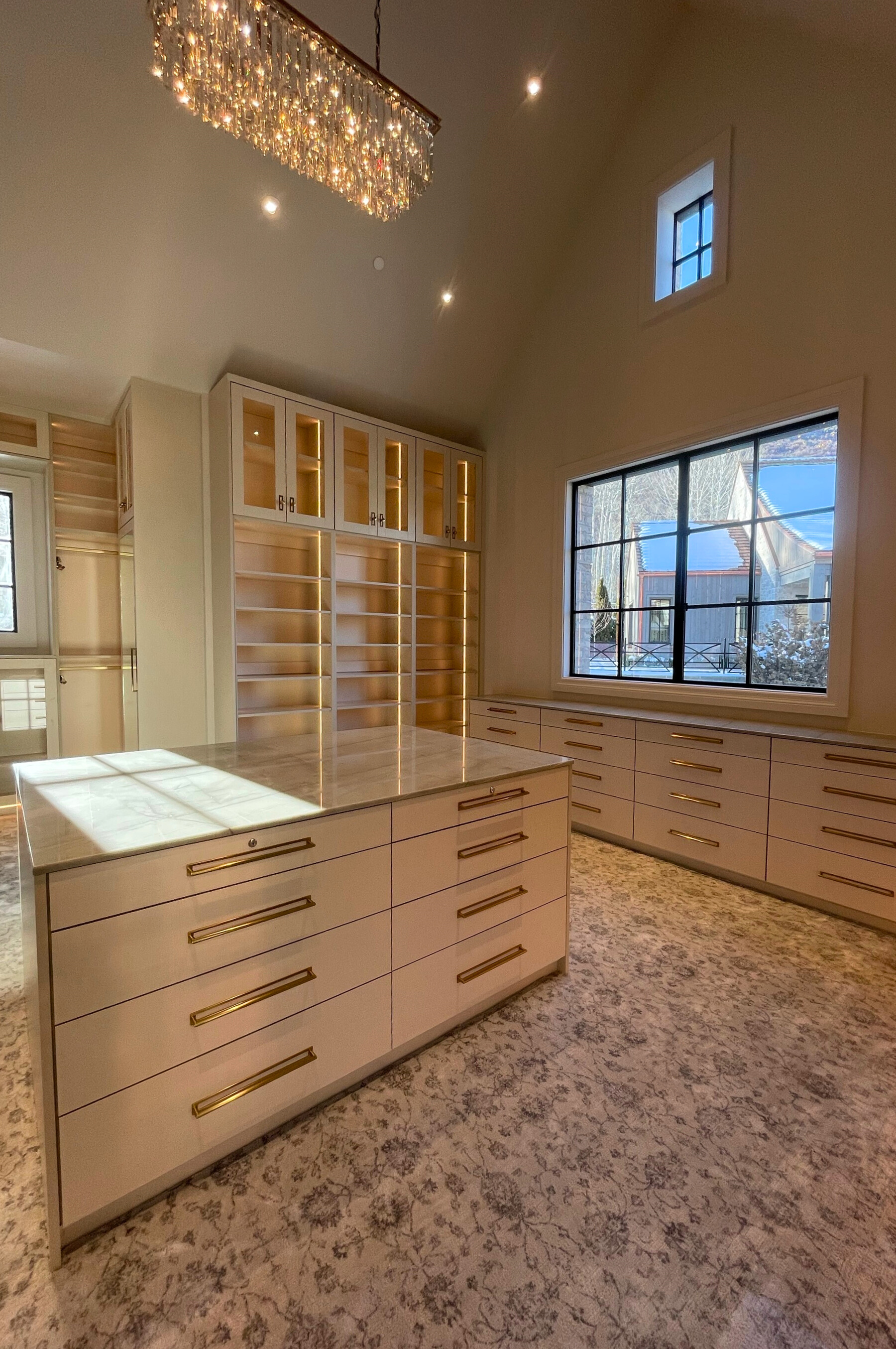 Luxury walk-in closet with built-in cabinets, a central island, and large windows. Neutral tones, chandelier.