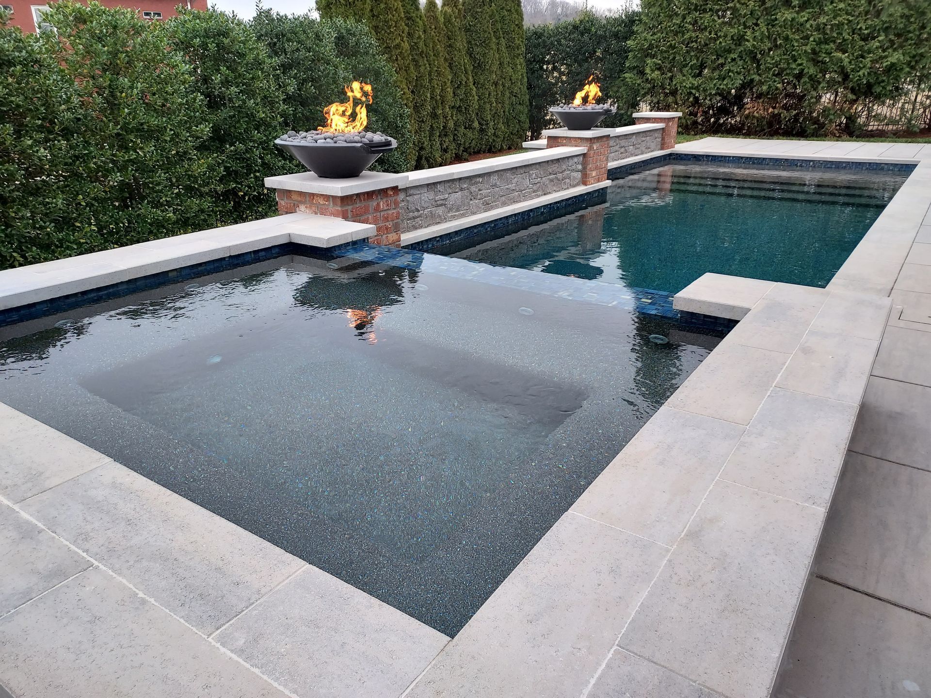 Pool with built-in spa, two fire bowls, surrounded by light gray stone patio and green hedges.