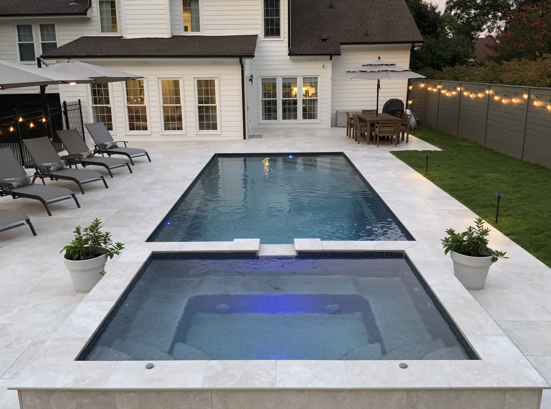 Backyard with a rectangular pool and hot tub, surrounded by light-colored stone patio.
