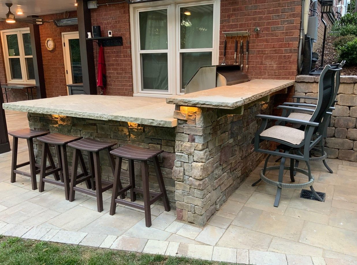 Outdoor bar with stone facade, counter, and stools. Built-in grill, patio setting with grass.