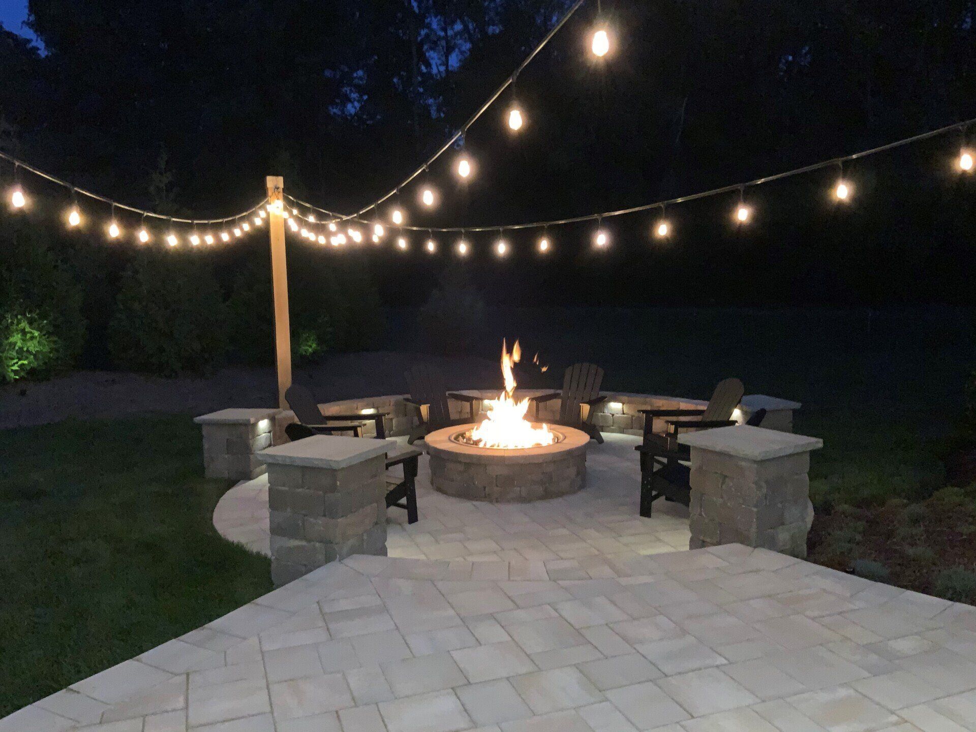 Lit backyard patio with fire pit and string lights at night.