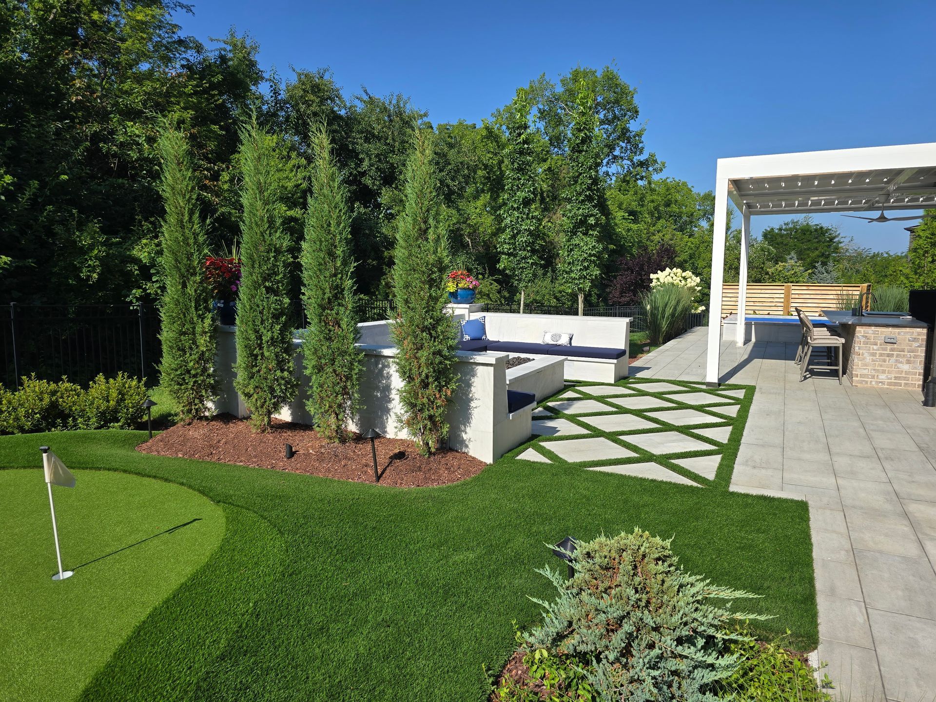 Modern landscaped backyard with green lawn, tall trees, stone patio, and white pergola