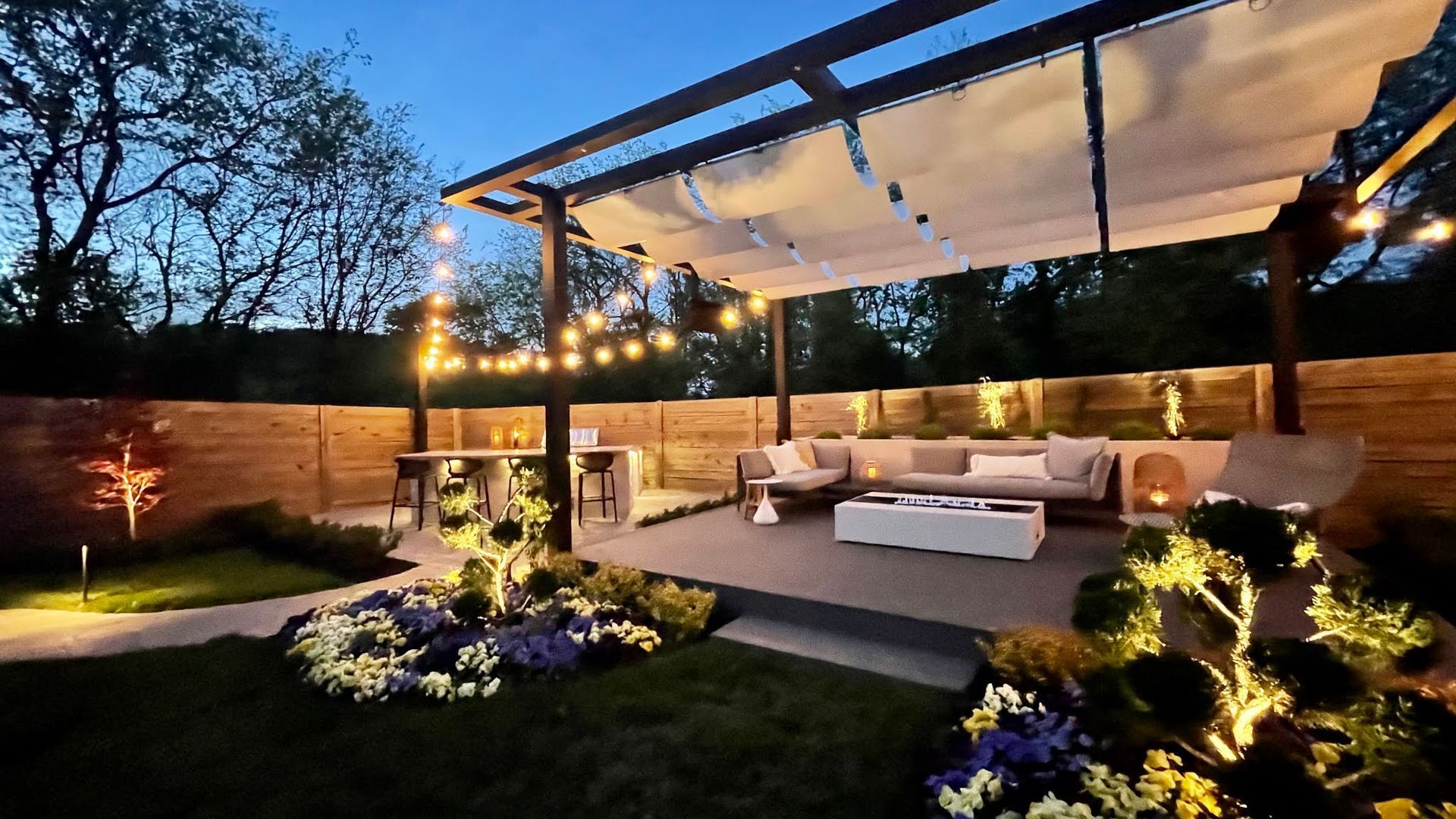 Outdoor patio at night with string lights, pergola, seating area, and landscaping.