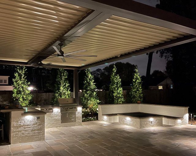 Nighttime patio with overhead structure, built-in grill, seating, and decorative trees, illuminated by warm lighting.