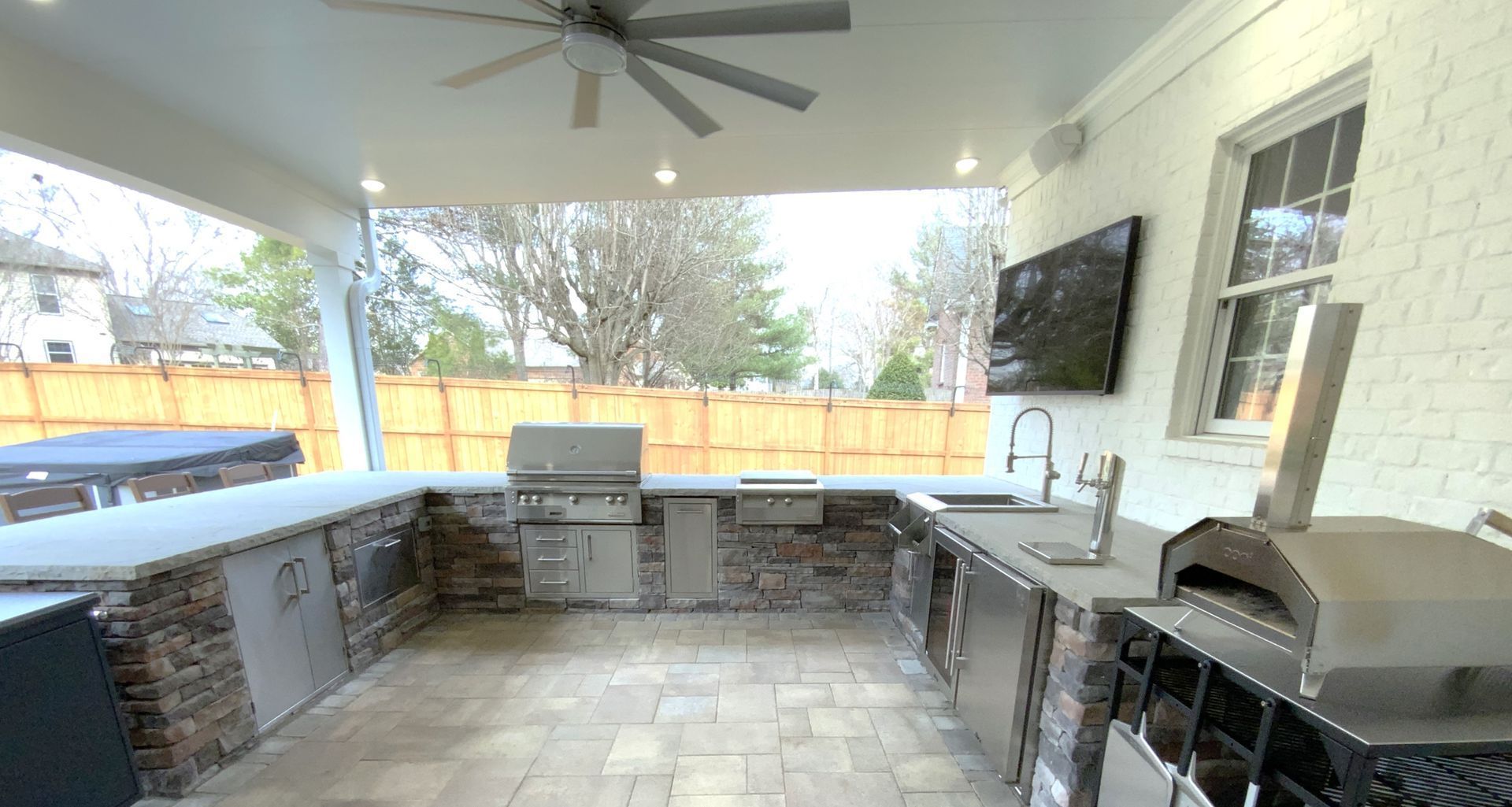 Outdoor kitchen with stone and stainless steel surfaces, grill, sink, and pizza oven under a covered patio.