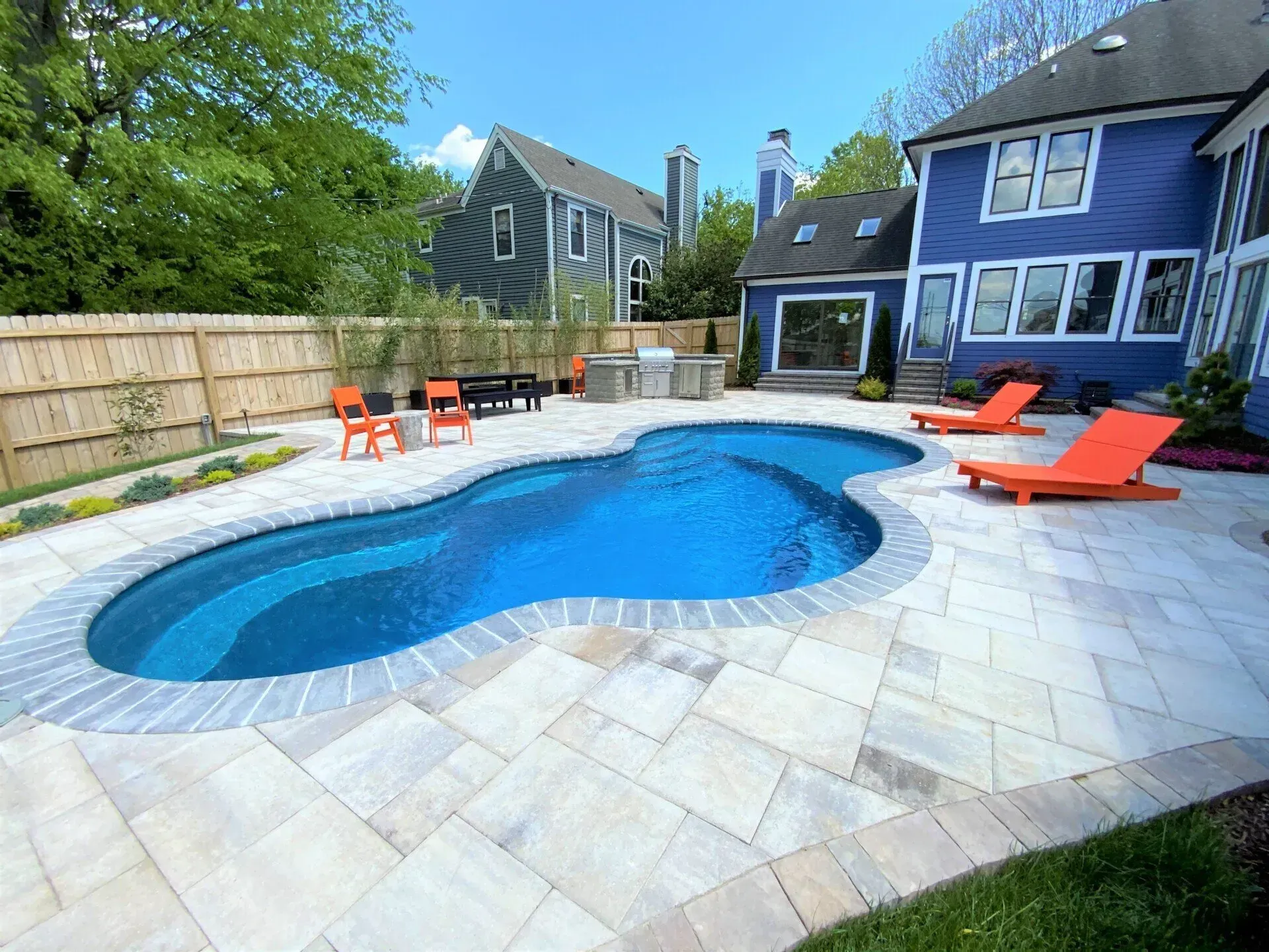 Backyard pool with light-colored pavers, surrounded by landscaping, next to a blue house with lounge chairs.