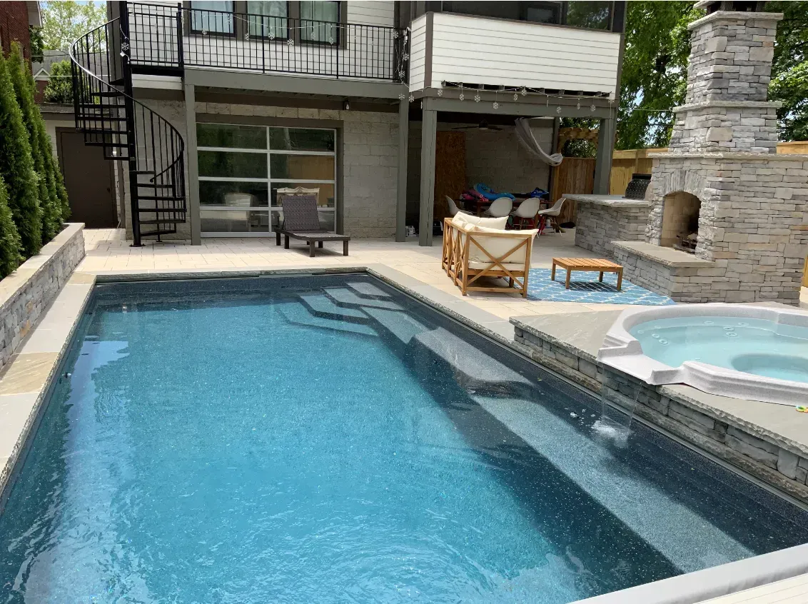 Backyard pool with steps, hot tub, fireplace, and spiral staircase on a sunny day.