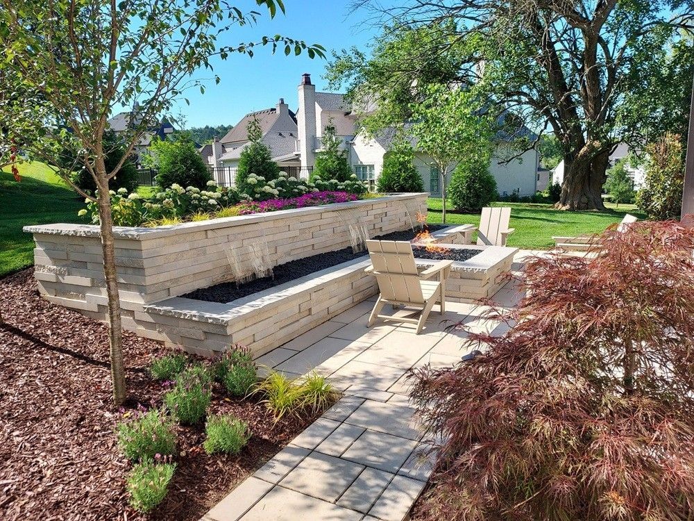 A landscaped backyard with a tiered stone wall water feature, patio, fire pit, and lounge chairs.