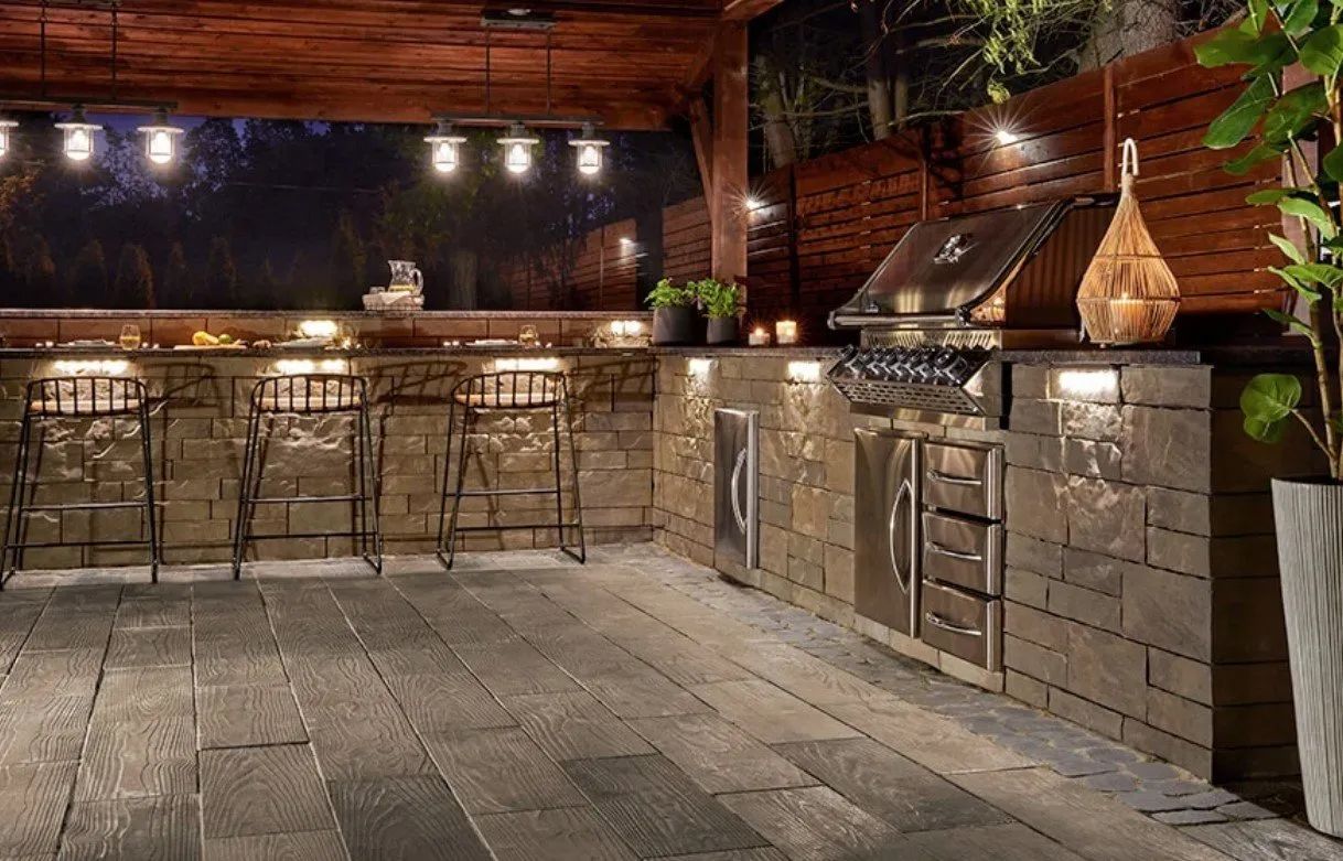 Outdoor kitchen at night with bar seating, grill, lighting, and stone facade.