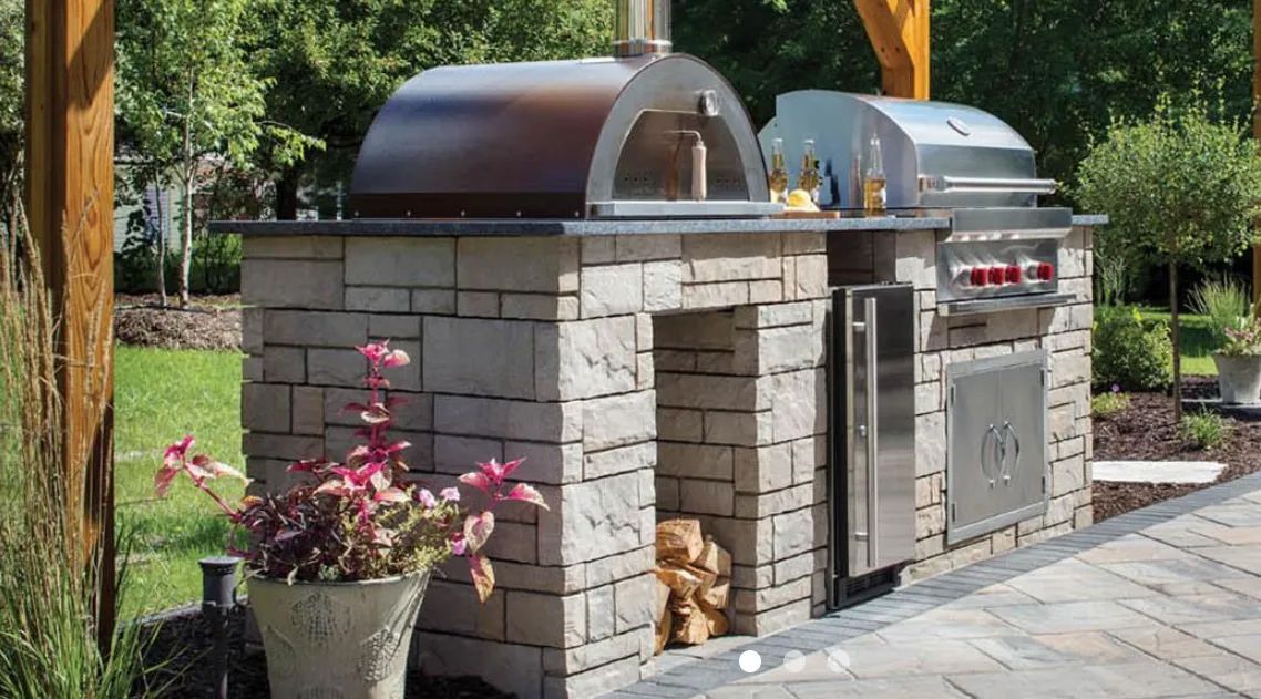 Outdoor kitchen with stone base, pizza oven, grill, and refrigerator.