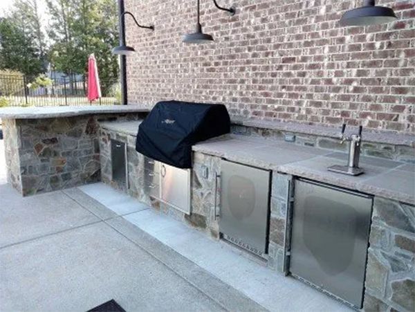 Outdoor kitchen with stone and stainless steel appliances, grill, countertop, brick wall, and pendant lights.