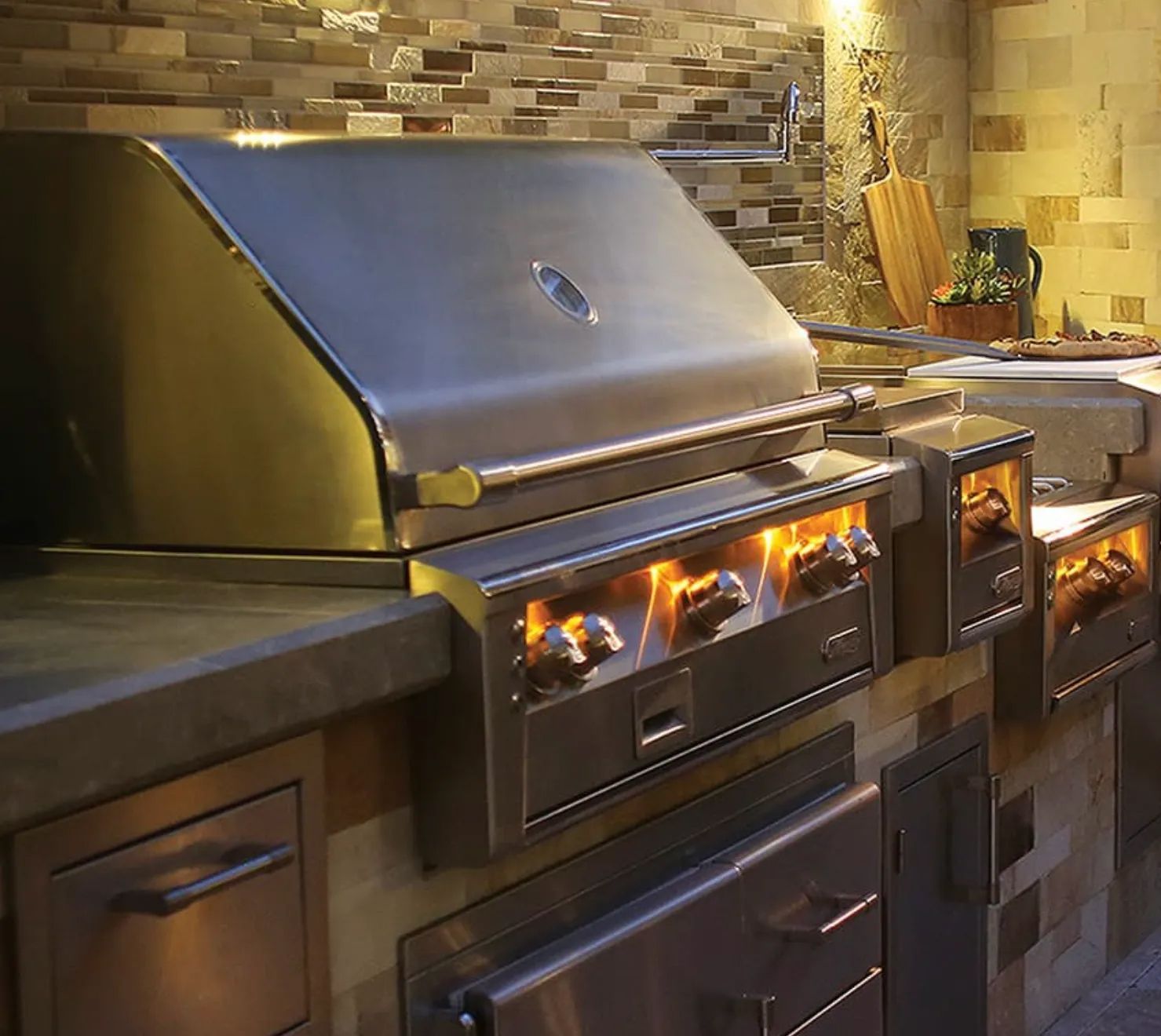Stainless steel outdoor grill with lit burners built into a stone and concrete countertop.