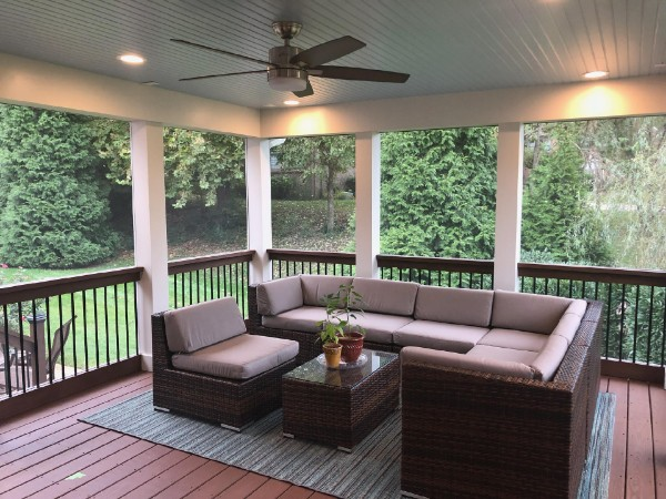 Screened-in porch with brown wicker furniture, ceiling fan, recessed lights, and views of greenery.
