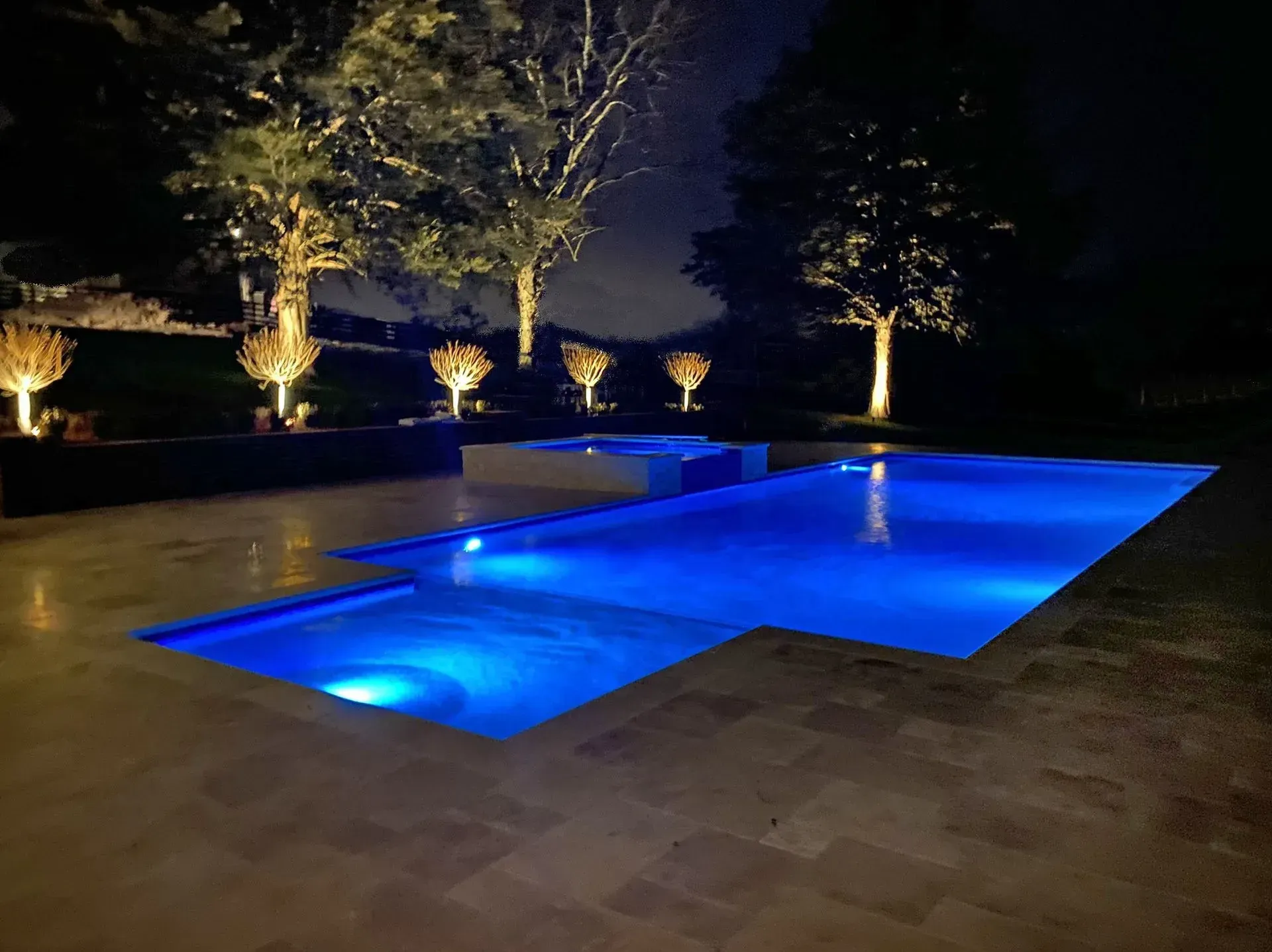 Nighttime view of a pool with blue underwater lights, trees with spotlights, and a dark sky.