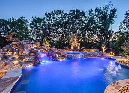 Night view of a luxury pool with waterfall, stone features, and fireplace, illuminated by blue and warm lights.