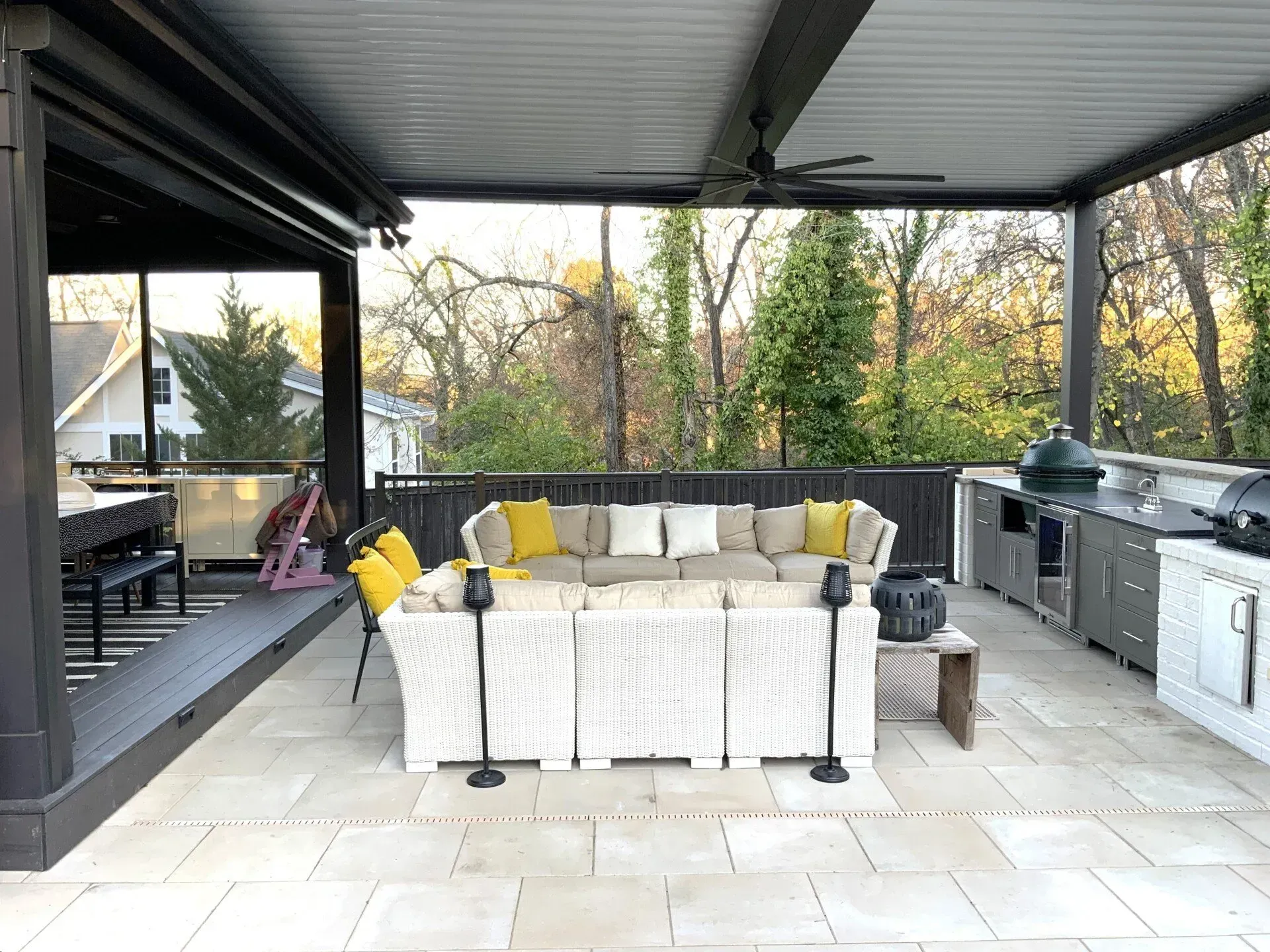 Outdoor patio with white woven sectional, outdoor kitchen, and overhead fan, with a view of trees.