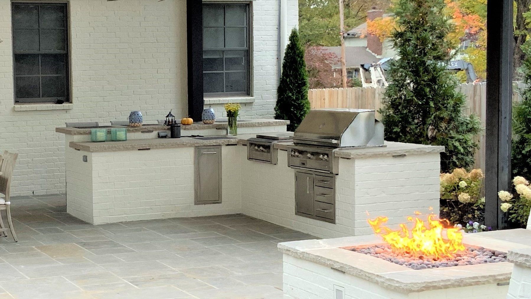 Outdoor kitchen with grill, sink, and fire pit on a patio, with brick wall and trees.