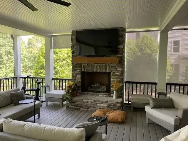Screened porch with fireplace, gray furniture, and roll-down shades. Overlooks a lush green treeline.