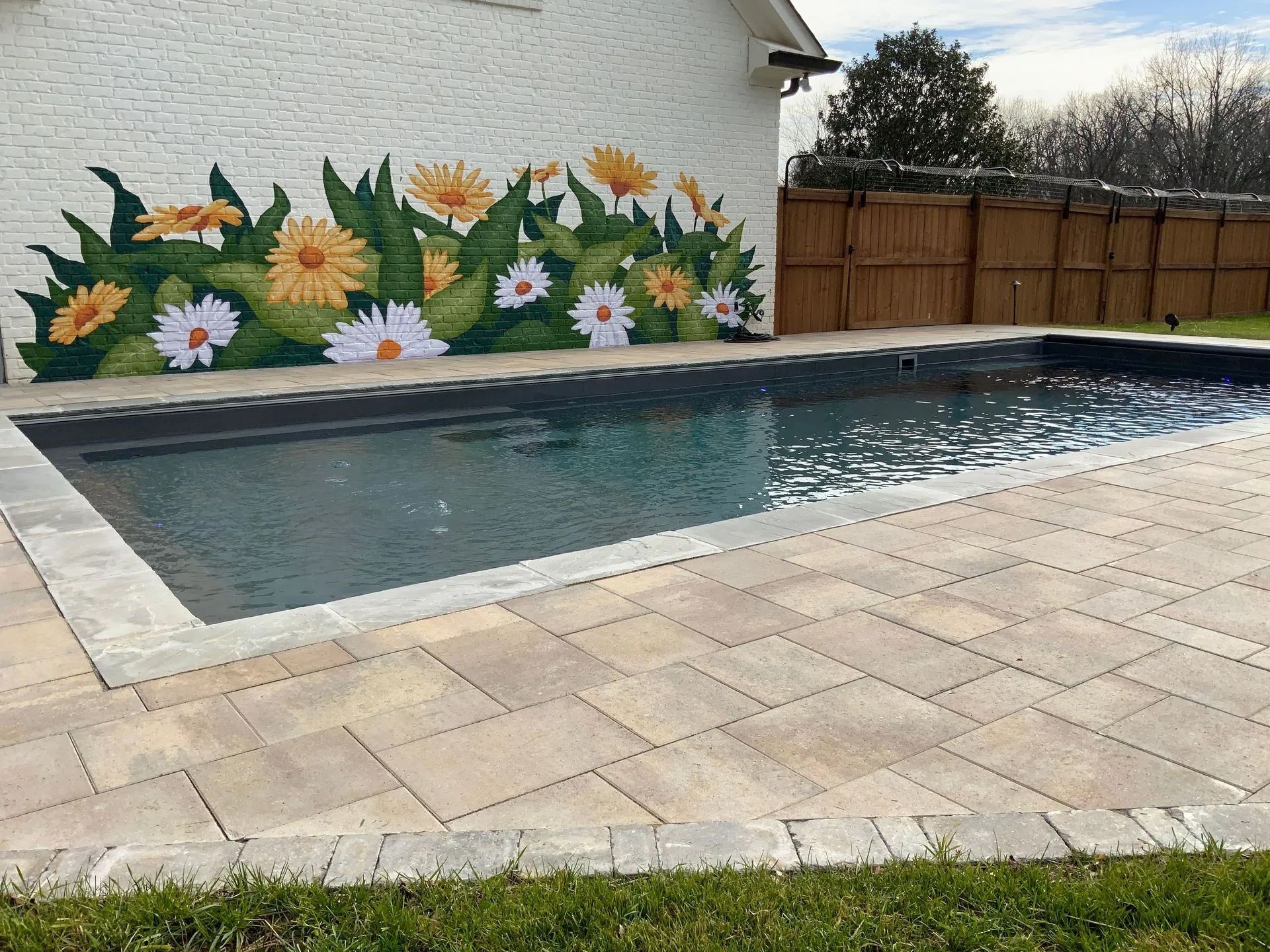 Swimming pool with daisy mural on the white brick wall, surrounded by stone patio and green grass.