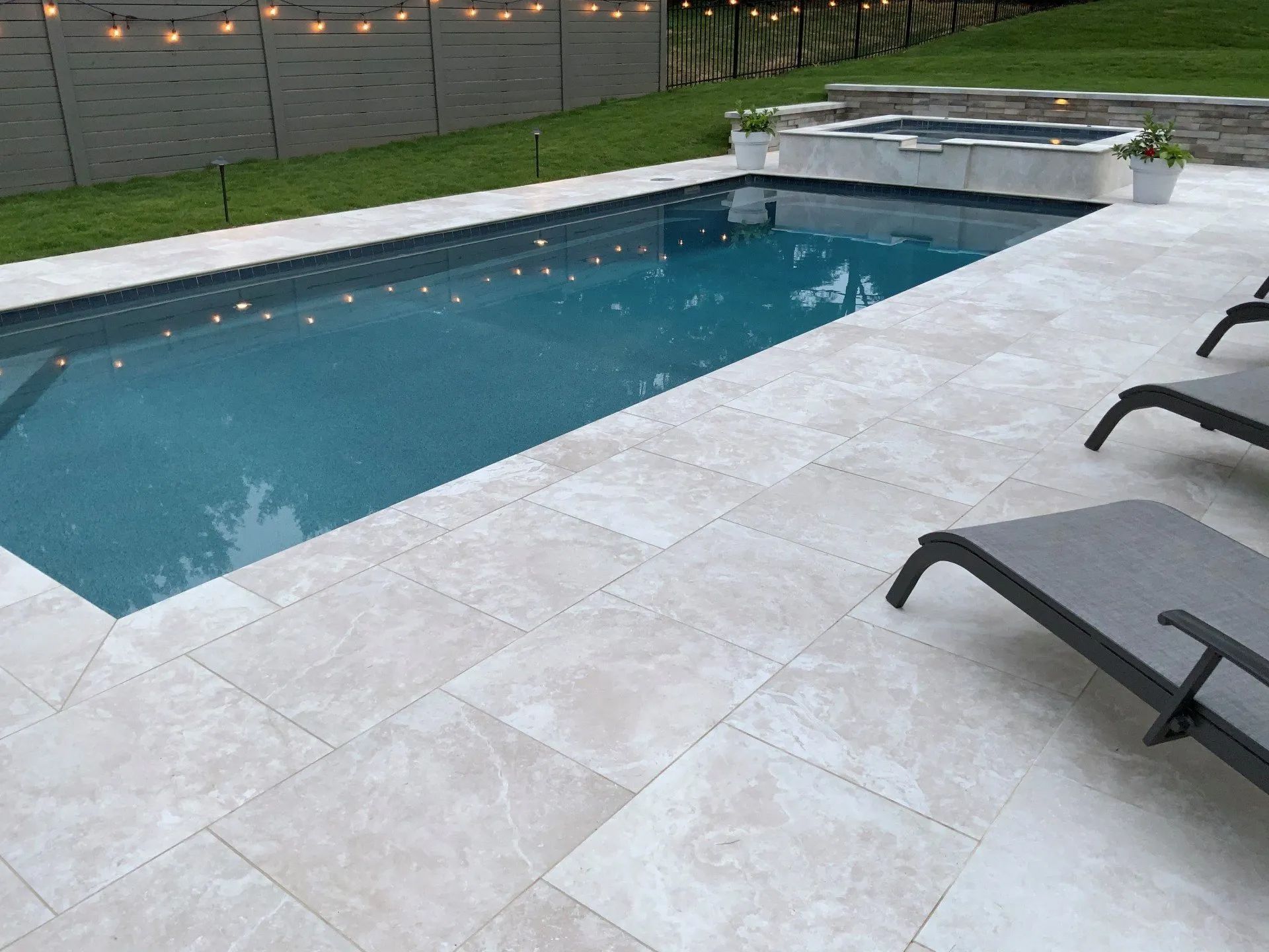 Swimming pool with light-colored stone patio, lounge chairs, and a small hot tub.