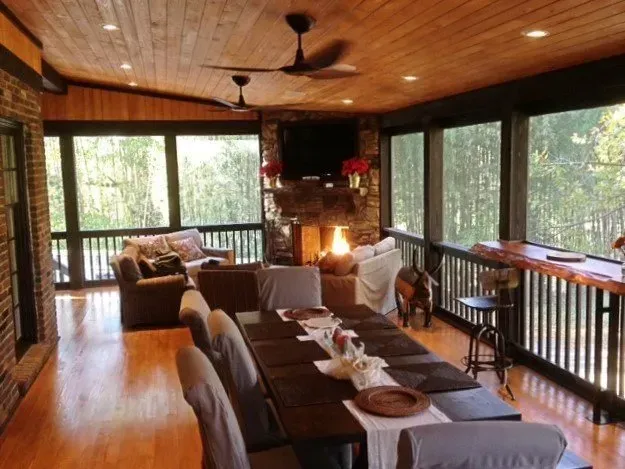 Cozy screened porch with dining table, fireplace, and seating, overlooking a wooded view.